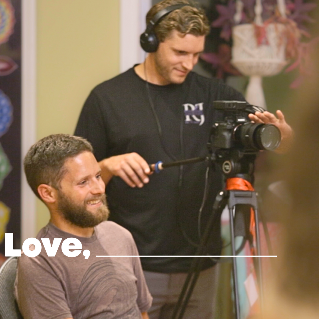 Two men, one standing and one sitting, look at a camera with lights for filming.